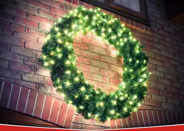 Close up view of a green wreath with warm white LED lights hung on a red brick exterior wall above a residential garage.